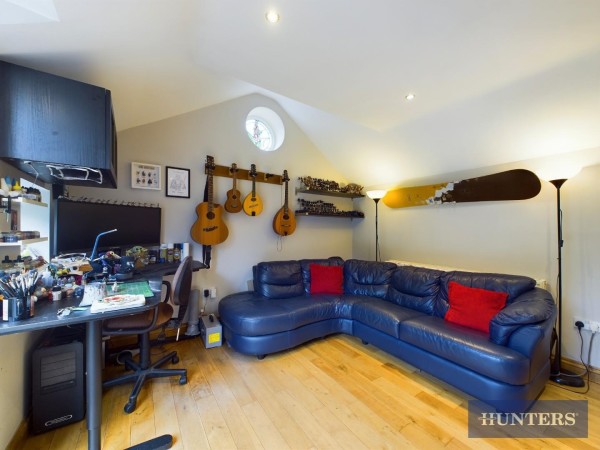 An annexe currently set up as a home office. There's a desk to the left and a black leather corner sofa with red cushions. Guitars hang on the walls and the floor is wooden.