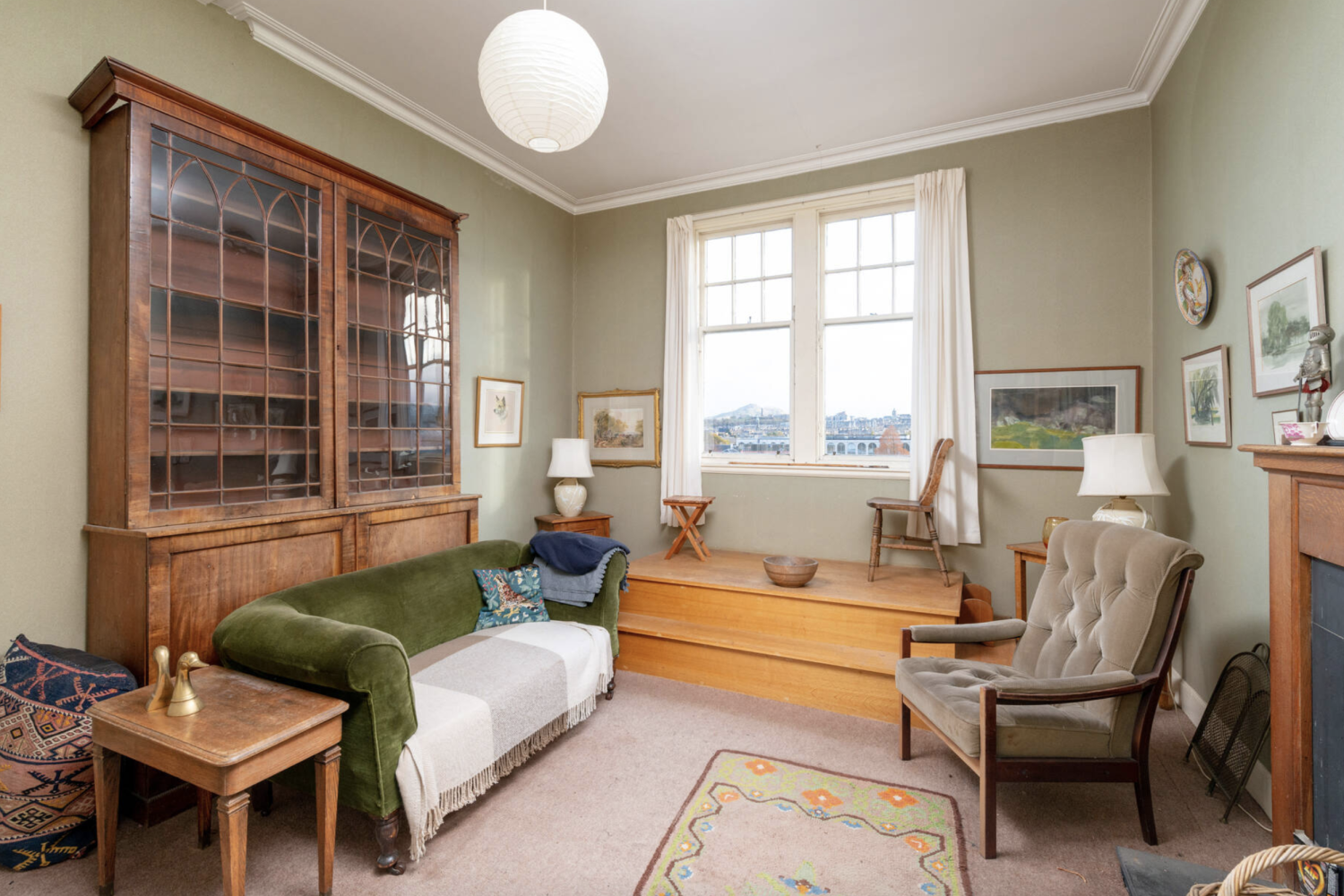 Interior of a Victorian apartment living room with pale green walls, a dresser, sofa and armchair and a viewing platform with chairs at the window