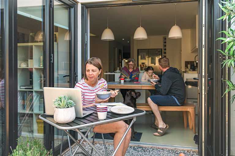 A woman sat at a table looking at her laptop in the garden with her family behind her in the house