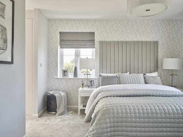 A corner of a stylish grey-toned bedroom, with a plush double bed with tall headboard, small window with grey blinds, and bedside tables with white lamps