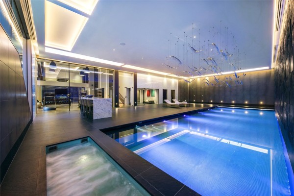 A large indoor pool with modern lighting and dark grey colour scheme on the surrounding walls and tiled flooring. There are sun beds and a bar around the edge and you can see a gym through glass doors.