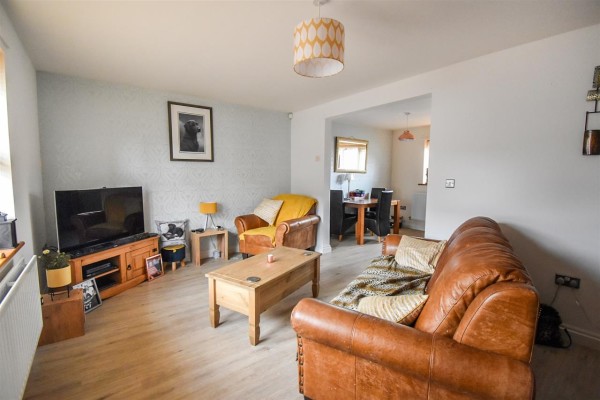 The lounge of a three bed end terrace house in Maryport. There is neutral decor with wooden floors, grey walls and a matching tan sofa and armchair pointing towards the TV in the corner. An archway leads to a dining area in the rear.