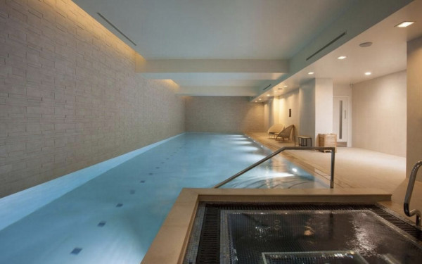 Beautiful blue indoor pool in an apartment complex, with a jacuzzi in the foreground and lounging area to one side, with the interior all fitted in pale stone