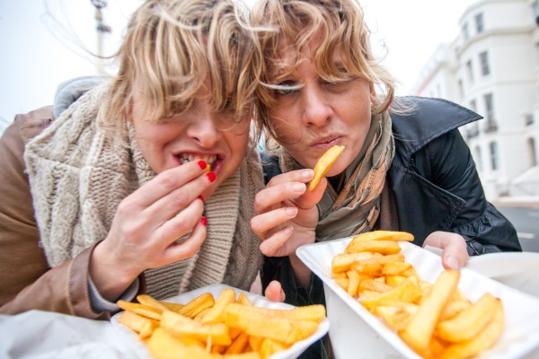 Two friends sharing a box of chips by the seaside on a windy day