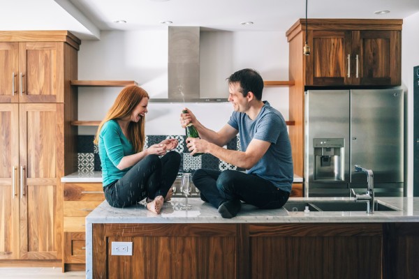 A couple cracking open a bottle of champagne while sitting on kitchen worktops