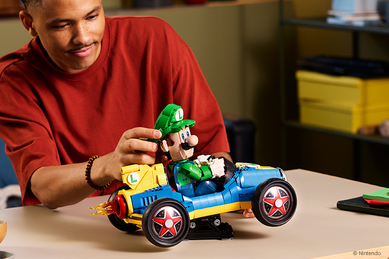 A man in a red shirt smiles while assembling a LEGO model of a blue and yellow racing car with a Luigi figure seated inside.