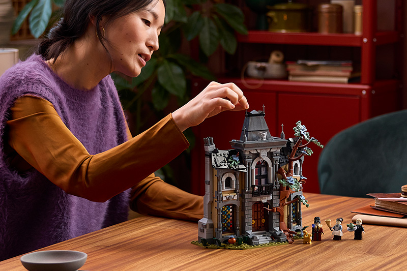 an adult building a detailed, gothic-style LEGO building set at a wooden table, with several minifigures displayed beside it. A red “NEW” badge appears in the upper-left corner.