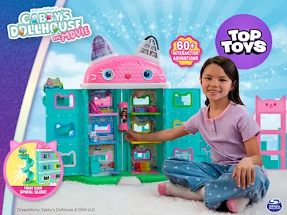 Girl sitting beside the Gabby's Dollhouse The Movie Interactive Playset Meow-mazing Dollhouse on a white carpeted floor of a bedroom.