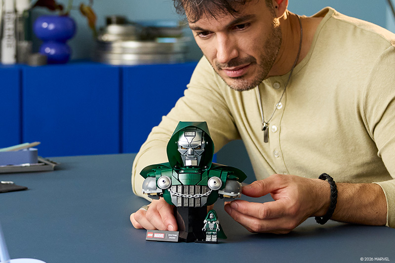"NEW" badge on a LEGO Marvel Doctor Doom bust set with a small minifigure, displayed on a table as a man in a tan shirt looks closely at it.