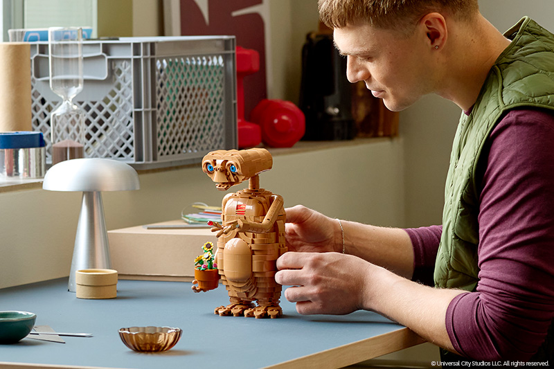 A person assembles a LEGO Ideas E.T. the Extra-Terrestrial model at a table. The detailed brick-built E.T. figure holds a small pot of flowers while the builder carefully adjusts the model in a bright indoor workspace.