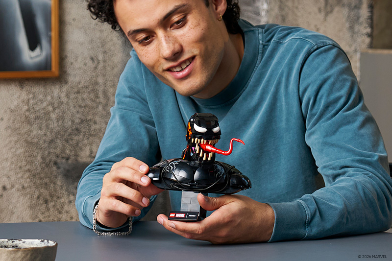 A person in a blue sweatshirt smiles while holding and adjusting a detailed brick-built bust of Venom on a display stand, featuring large teeth, white eyes, and an extended red tongue, on a tabletop indoors.