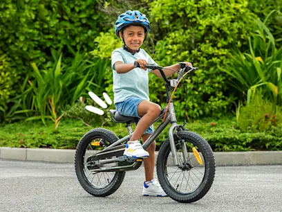Smiling child in a blue helmet rides a bike on a paved path, surrounded by green foliage.