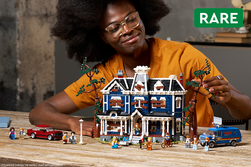 A woman examines a detailed LEGO model of the Creel House from Stranger Things, surrounded by trees, minifigures, vehicles and accessories on a wooden table. A green “RARE” label appears in the upper right.