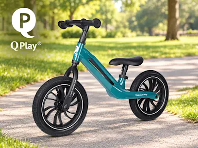 Q Play balance bike with a teal frame, black seat, handlebars and two large black wheels, pictured on a paved path in a sunny green park.