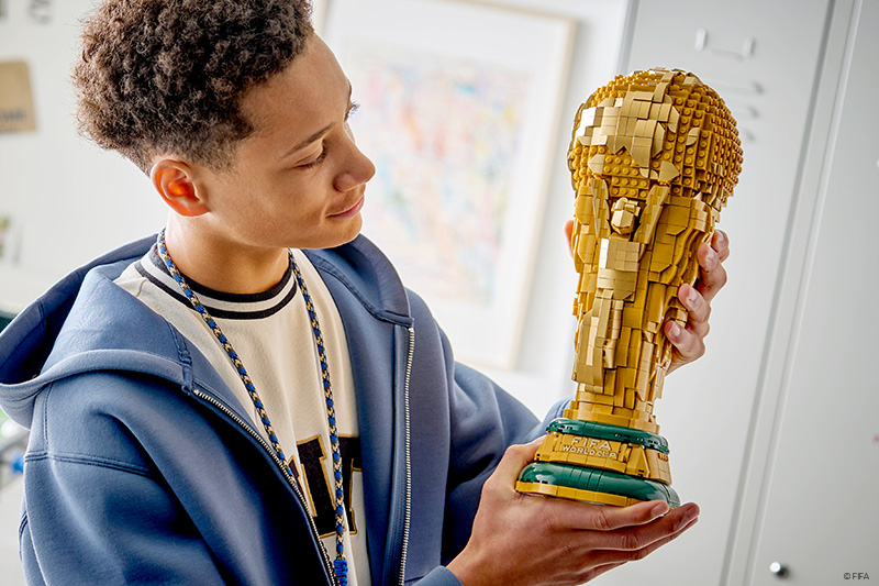 A child in a blue hoodie holds and examines a detailed brick-built replica of the FIFA World Cup Trophy indoors, looking closely at the gold-colored model while supporting its green base with both hands.