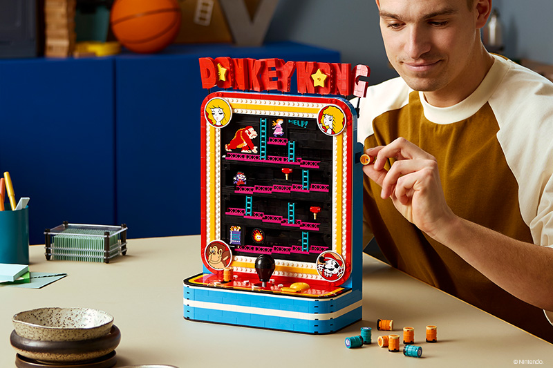 LEGO Donkey Kong arcade machine build on a desk, with a man placing small colored piece elements beside it as he assembles the model.