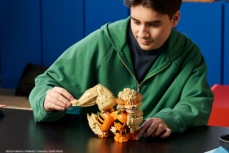 A child in a green hoodie builds a LEGO Pokémon Arcanine model at a table, carefully attaching its large tail. The orange-and-tan brick-built Pokémon is partially assembled in an indoor play setting.