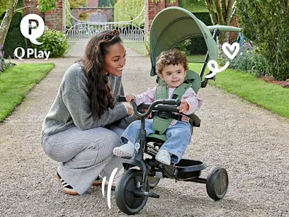Adult kneels beside a smiling toddler seated on a green tricycle stroller with a canopy, on a garden path. Decorative heart graphics and the Q Play logo appear, with a gated garden entrance in the background.