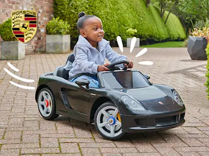 Child smiles while driving a black Porsche-style ride-on toy car along a paved garden path. White motion graphics and a Porsche logo appear on the image, with neatly trimmed hedges and planters in the background.