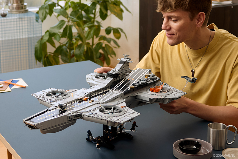 Man in a yellow shirt happily assembles a detailed LEGO model of a large aircraft carrier on a blue table. A plant and mug are in the background.