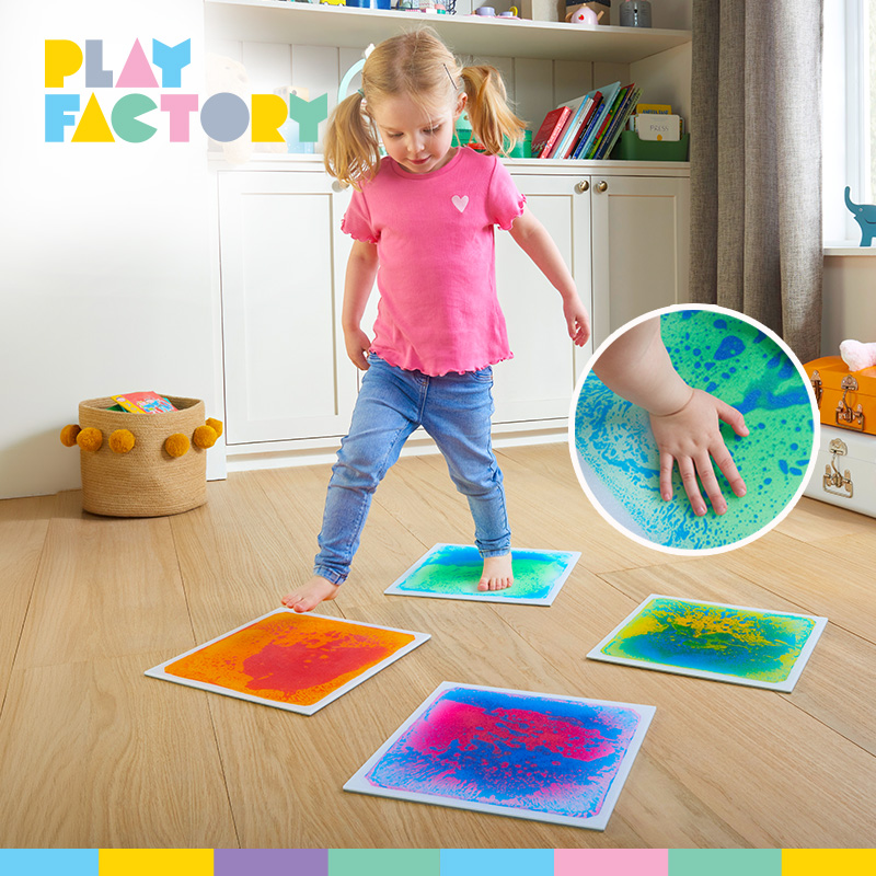Play Factory activity showing a young girl stepping on colourful sensory mats that create patterns underfoot. Four textured mats in blue, orange, green and pink are spread across a wooden floor.