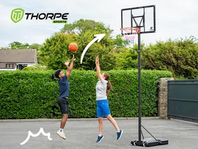 Two people playing basketball outdoors in a driveway, one shooting a ball toward a hoop. Lush greenery and a basketball stand in the background.