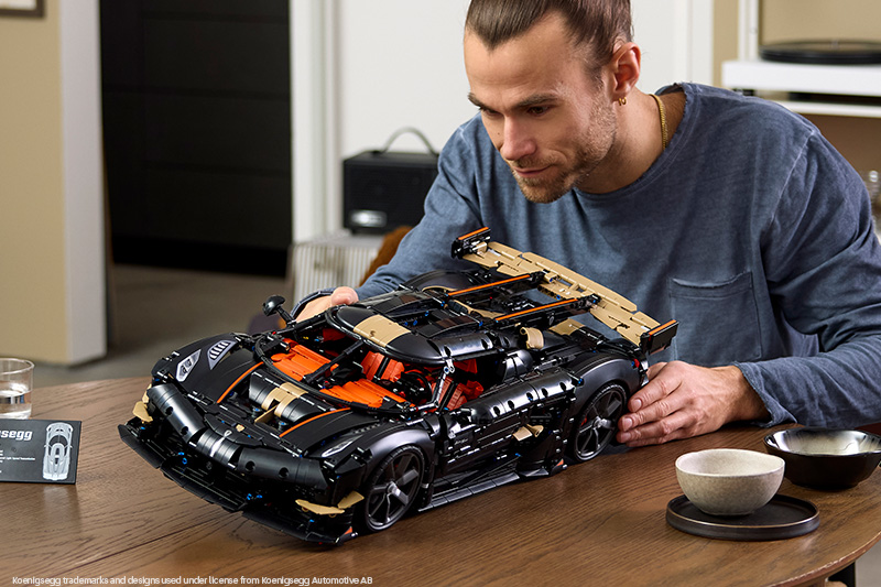 LEGO Technic Koenigsegg supercar set displayed on a table, with a man examining the detailed black, orange and gold model. The image highlights the car’s realistic design and intricate construction.