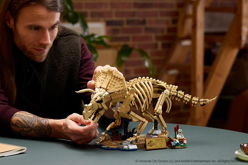 A person with long hair and a tattoo inspects a detailed LEGO triceratops skeleton model on a table.