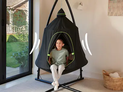Child sits smiling inside a black sensory swing with an enclosed cocoon-style seat on a freestanding frame indoors. White motion lines suggest gentle swinging, with a bright room and garden visible through a nearby glass door.