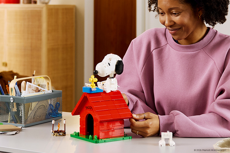 A person in a pink sweatshirt smiles while assembling a brick-built scene featuring a white cartoon dog on a red doghouse, adjusting the roof with both hands at a desk with craft supplies nearby.