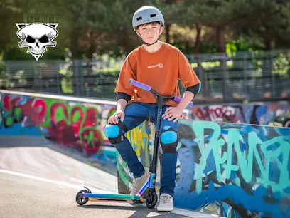 Child wearing a helmet, knee pads, and elbow pads sits on a stunt scooter at a skate park, with colorful graffiti-covered ramps in the background. A Blindside skull logo appears in the top left corner.