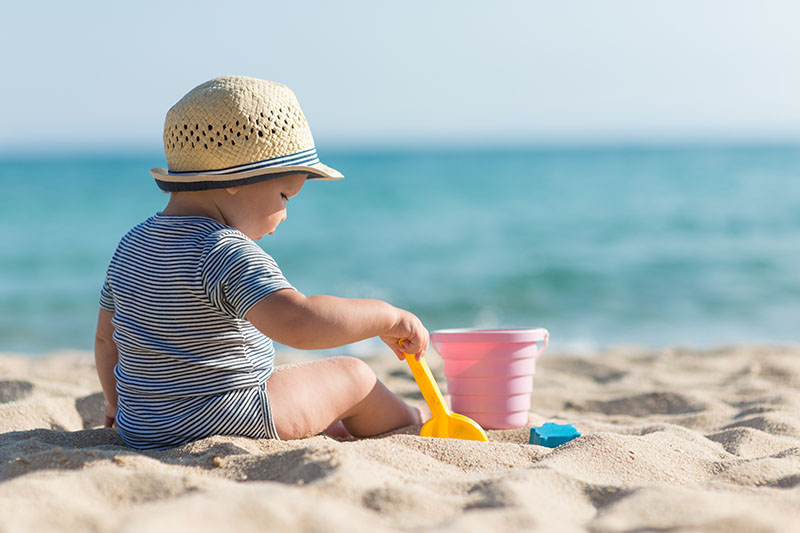A baby sitting on a sandy beach playing with beach toys.