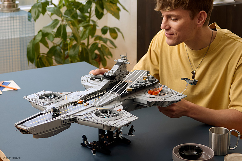 A person in a yellow shirt admires a large brick-built aircraft carrier model on a table, holding one side while looking at the detailed deck, aircraft, and display features in a bright indoor setting.