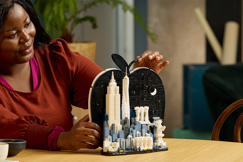 A person in a red sweater admires a detailed Lego model of New York City, featuring skyscrapers and the Statue of Liberty, shaped like an apple.