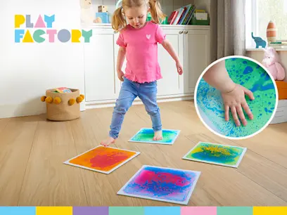 A young child presses their hands into four colorful sensory paint boards on the floor, creating green, purple, blue, pink, and yellow patterns. A Play Factory logo appears in the upper-left corner.