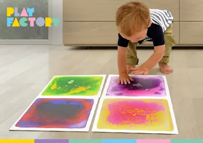 A young child presses their hands into four colorful sensory paint boards on the floor, creating green, purple, blue, pink, and yellow patterns. A Play Factory logo appears in the upper-left corner.