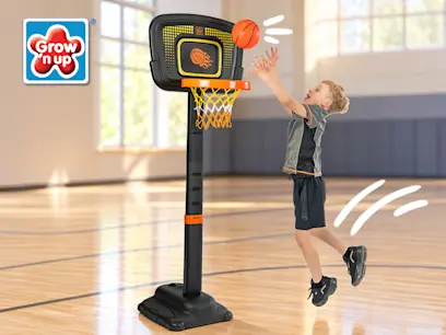 Grow’n Up adjustable basketball stand with a young child jumping to shoot a basketball. The black hoop has an orange rim and net and stands on a weighted base in an indoor basketball court.