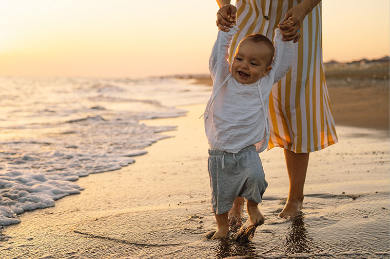 A Lady holding the hands of a baby as they walk together along the water's edge on a sandy beach.