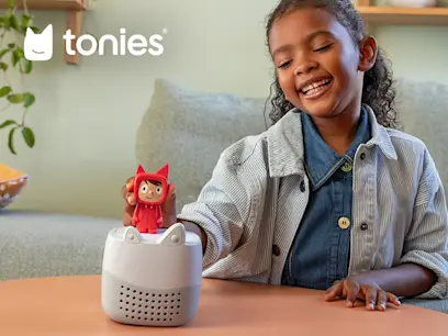 Tonies promotional image showing a smiling child placing a red Tonie character on a white Toniebox Lite audio player on a table, with the Tonies logo in the upper-left corner.