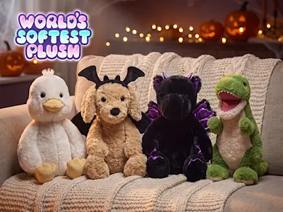 World’s Softest Plush Halloween display featuring four plush toys—a white duck, brown dog dressed as a bat, black bat, and green dinosaur—seated on a sofa with pumpkins and warm lights in the background.
