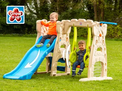 Two young children enjoy a beige plastic playset on green grass; one slides on a blue slide, the other swings on a lime green swing, both smiling.