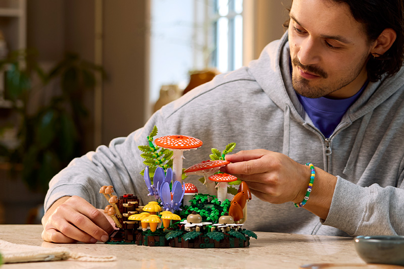 A person in a gray hoodie arranges a colorful brick-built woodland scene featuring mushrooms, plants, and small creatures on a table, carefully adjusting one of the red-capped mushrooms in a cozy indoor setting.