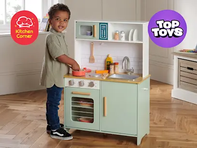 A kid standing beside and playing with the Kitchen Corner Wooden Play Kitchen in a room beside a window.