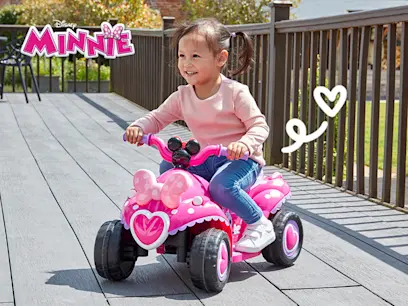 A young child rides a pink Minnie Mouse quad bike on a sunny wooden deck.