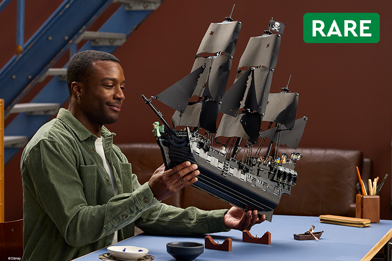 A man examines a detailed LEGO sailing ship model with black sails and pirate flags at a table. The model is displayed against a workshop-style background, with a green “RARE” label in the upper right.