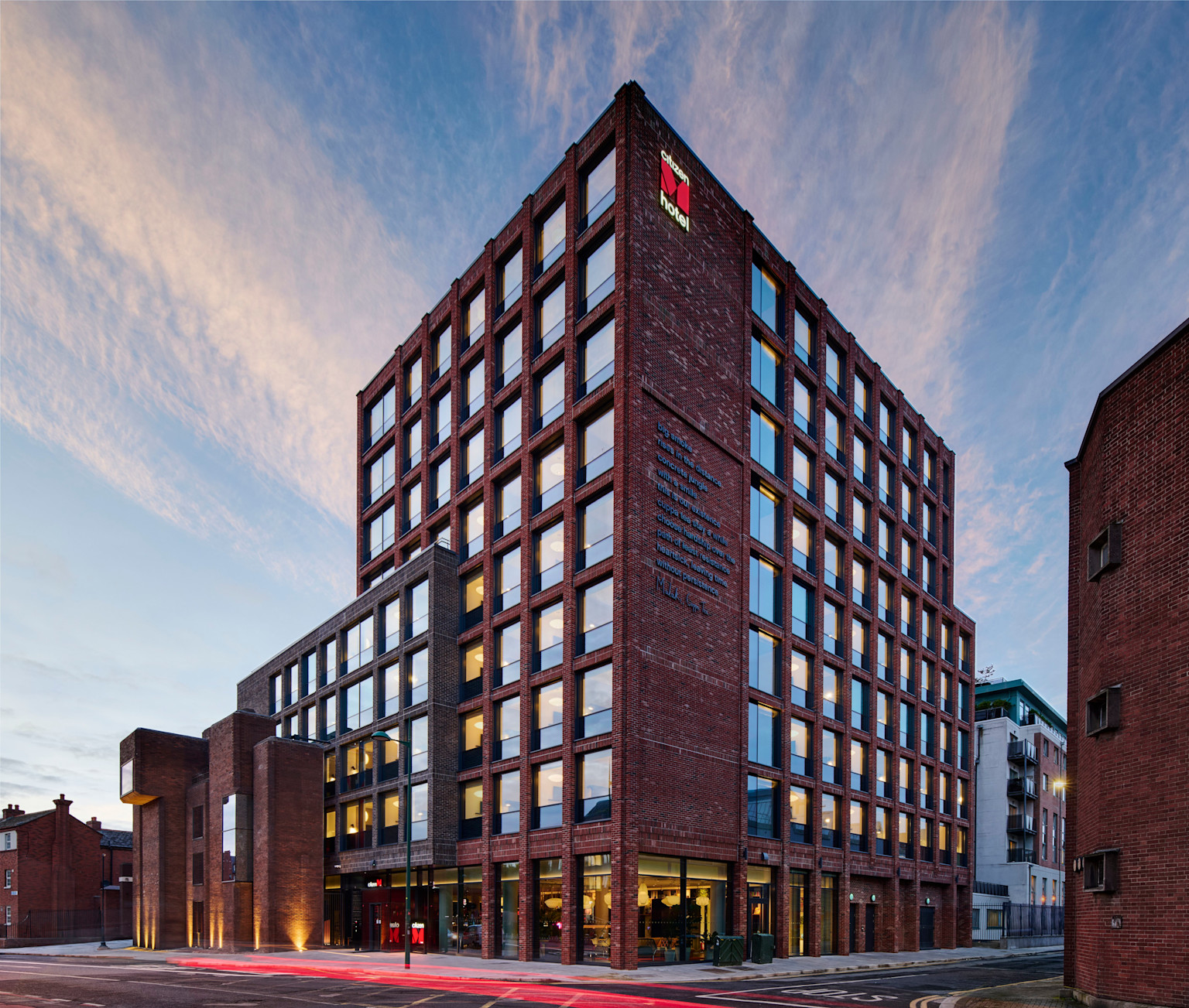 Exterior view of the citizenM Dublin St. Patrick’s hotel at dusk, showing its modern red-brick façade with large grid windows and warm interior lighting.