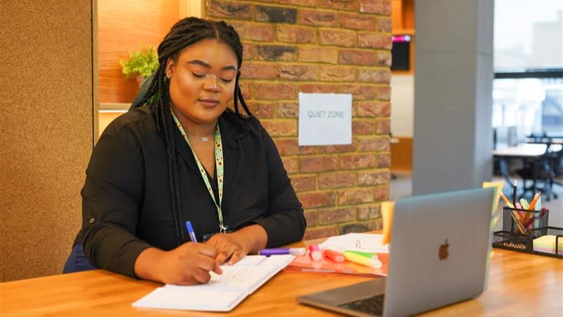 A woman working on a laptop, looking down, writing notes on a notepad, wearing an invisible disabilities sunflower lanyard.