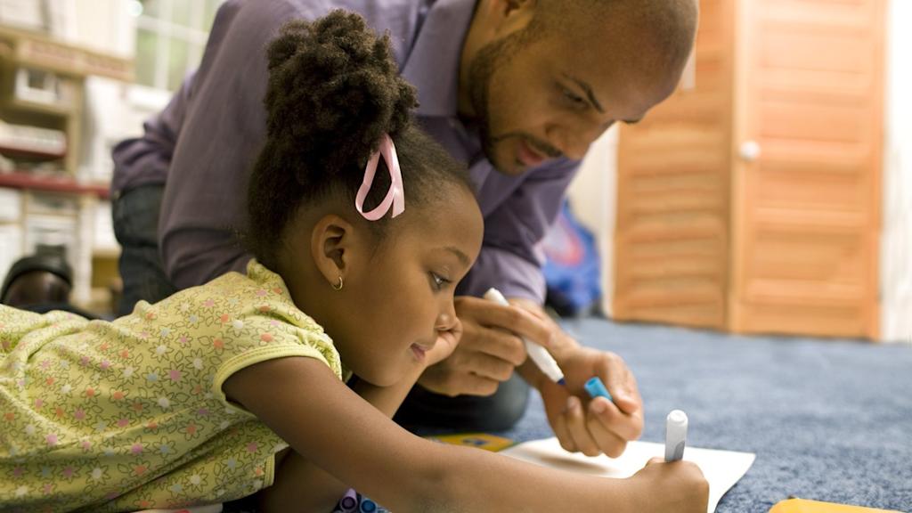A child lying down drawing on paper, with an adult crouched beside them also drawing.
