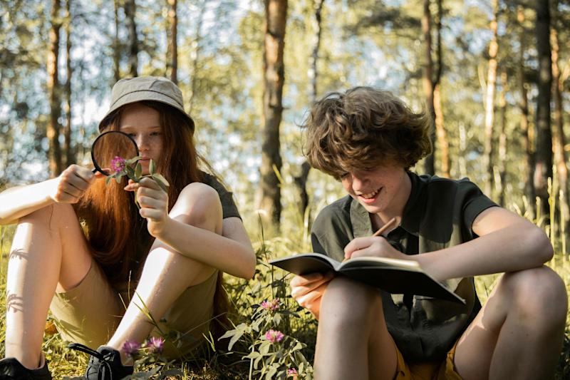 Two teenagers, around 13 years old, are sitting in a forest. In the background are rows of tall trees. The teenager on the left is looking at a pink flower with a magnifying glass, while the teenager on the right smiles and takes notes in an exercise book.
