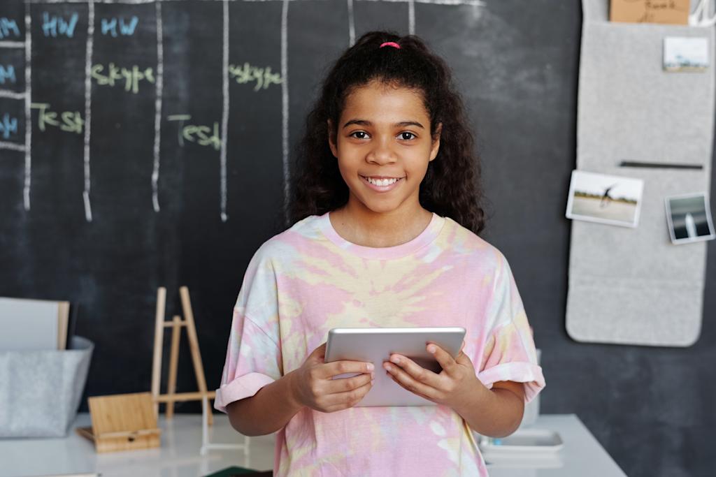 Smiling child looking directly at camera, holds tablet, with blackboard and table behind her